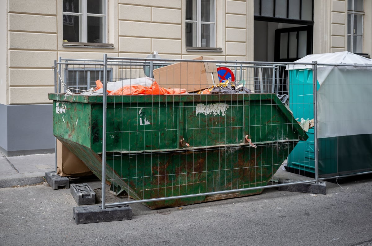Green dumpster full of construction waste behind metal fence on city street