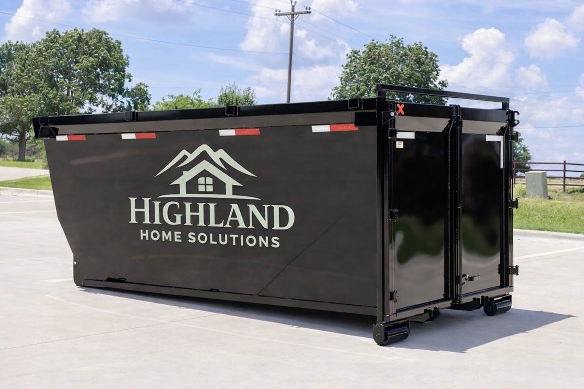 Black dumpster container with Highland Home Solutions logo and house icon parked in a rural setting with trees and power lines visible in the background